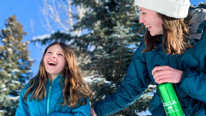 two girls laughing in the snow holding a water bottle