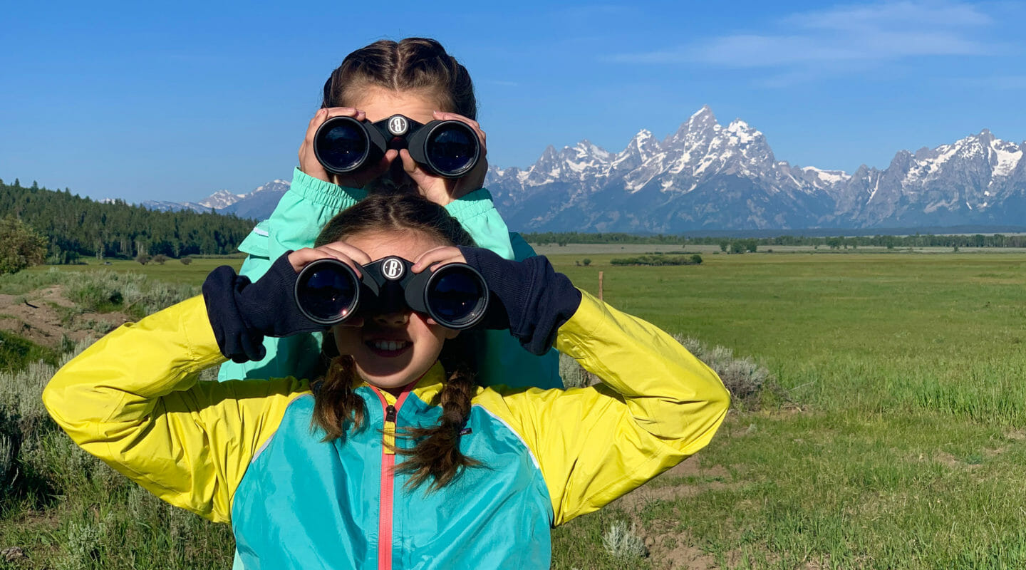 Two Girls Look For Wildlife Through Binoculars While On Safari In Grand Teton National Park With The Teton Mountain Range Visible Behind Them