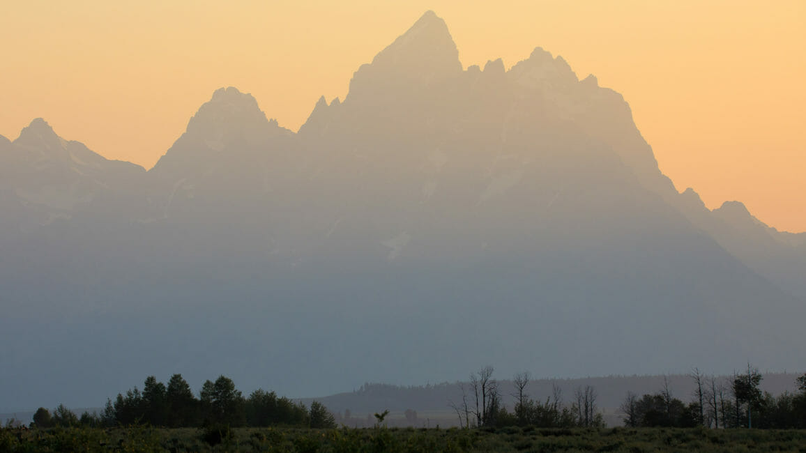 The Iconic Peaks Of The Teton Range Are Silhouetted Against A Warmly Lit Sunset In Grand Teton National Park
