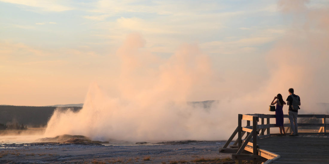 Two People Stand On The Boardwalk In Yellowstone National Park To Take A Photo Of Steam Rising From A Geyser