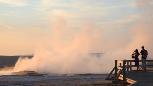 Two People Stand On The Boardwalk In Yellowstone National Park To Take A Photo Of Steam Rising From A Geyser
