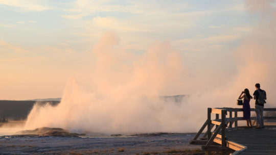 Two People Stand On The Boardwalk In Yellowstone National Park To Take A Photo Of Steam Rising From A Geyser